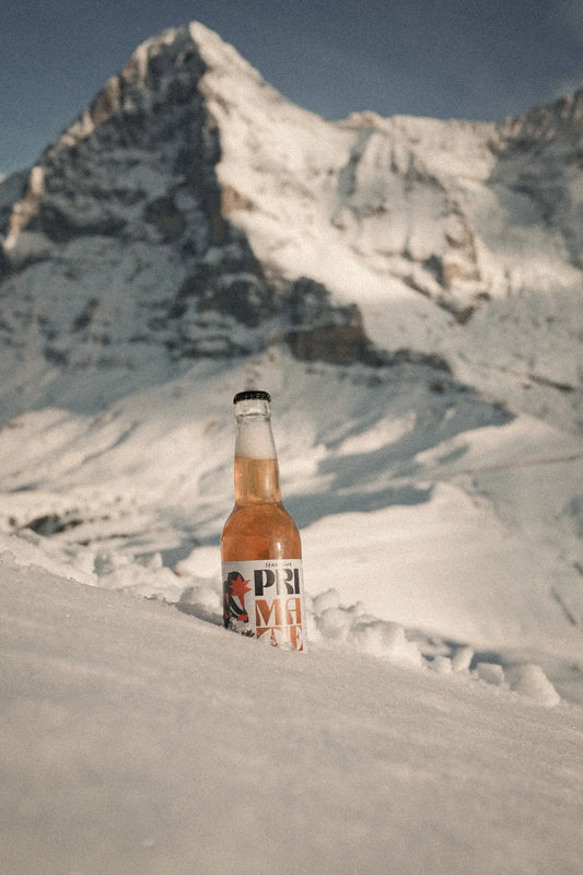 Flasche des Getränks PRI MATE Bio-Mate Sparkling steckt im Schnee vor einer verschneiten Berglandschaft mit sonnigem Himmel und einem markanten Gipfel im Hintergrund.