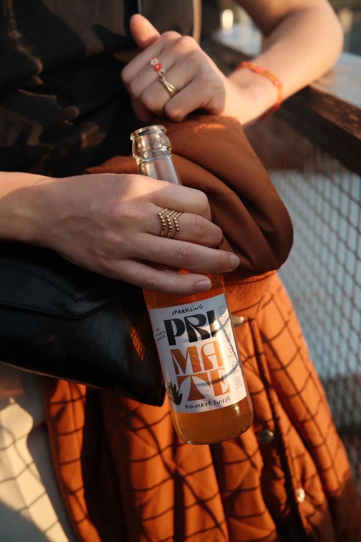Person hält eine Flasche PRI MATE Bio-Mate Sparkling in der Hand, im Sonnenlicht, mit Schmuckringen und orangefarbener Jacke im Hintergrund.